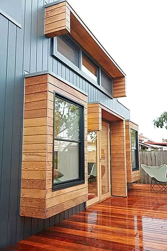 Timber-clad window boxes and entry on a dark vertical-board facade, opening onto an oiled hardwood deck