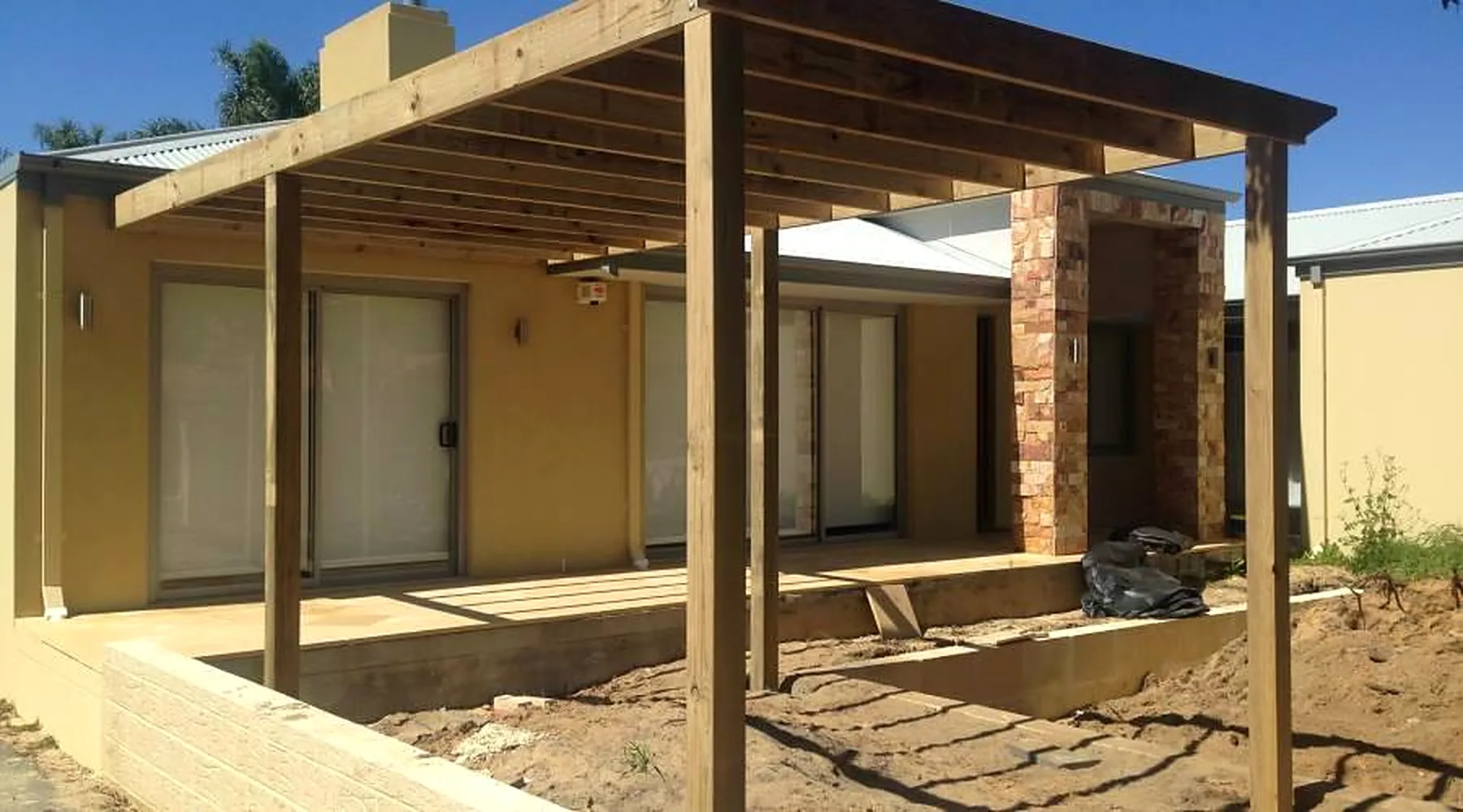 Large freestanding timber pergola frame over a new deck beside a stacked-stone pillar